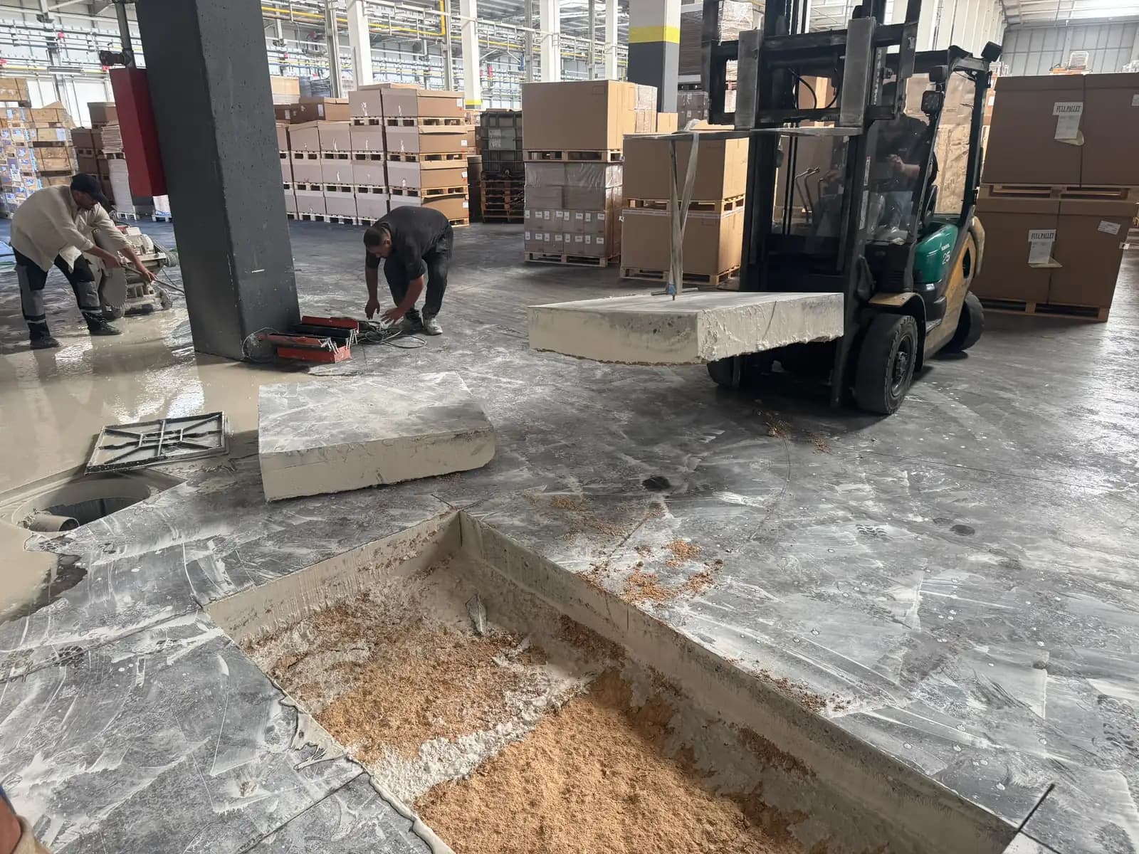 Forklift çatalına alınmış beton döşeme plakası; arkada paletli ürünler, önde talaşla doldurulmuş açıklık