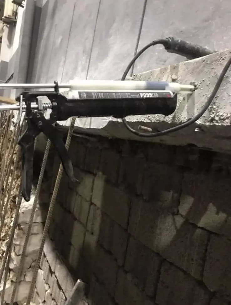 Beton yüzeyi üzerinde duran kimyasal ankraj tabancası ve arkada briket duvara bağlanan filiz donatıları
