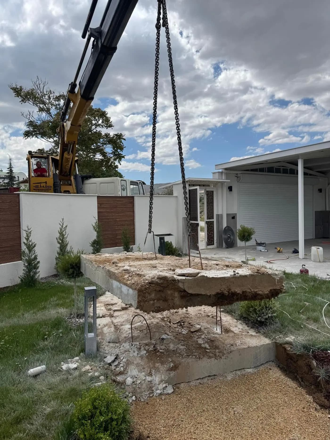 Peyzajlı villa bahçesinde vinç zinciriyle havaya kaldırılan beton plaka ve altında açığa çıkan dolgu ile donatılar