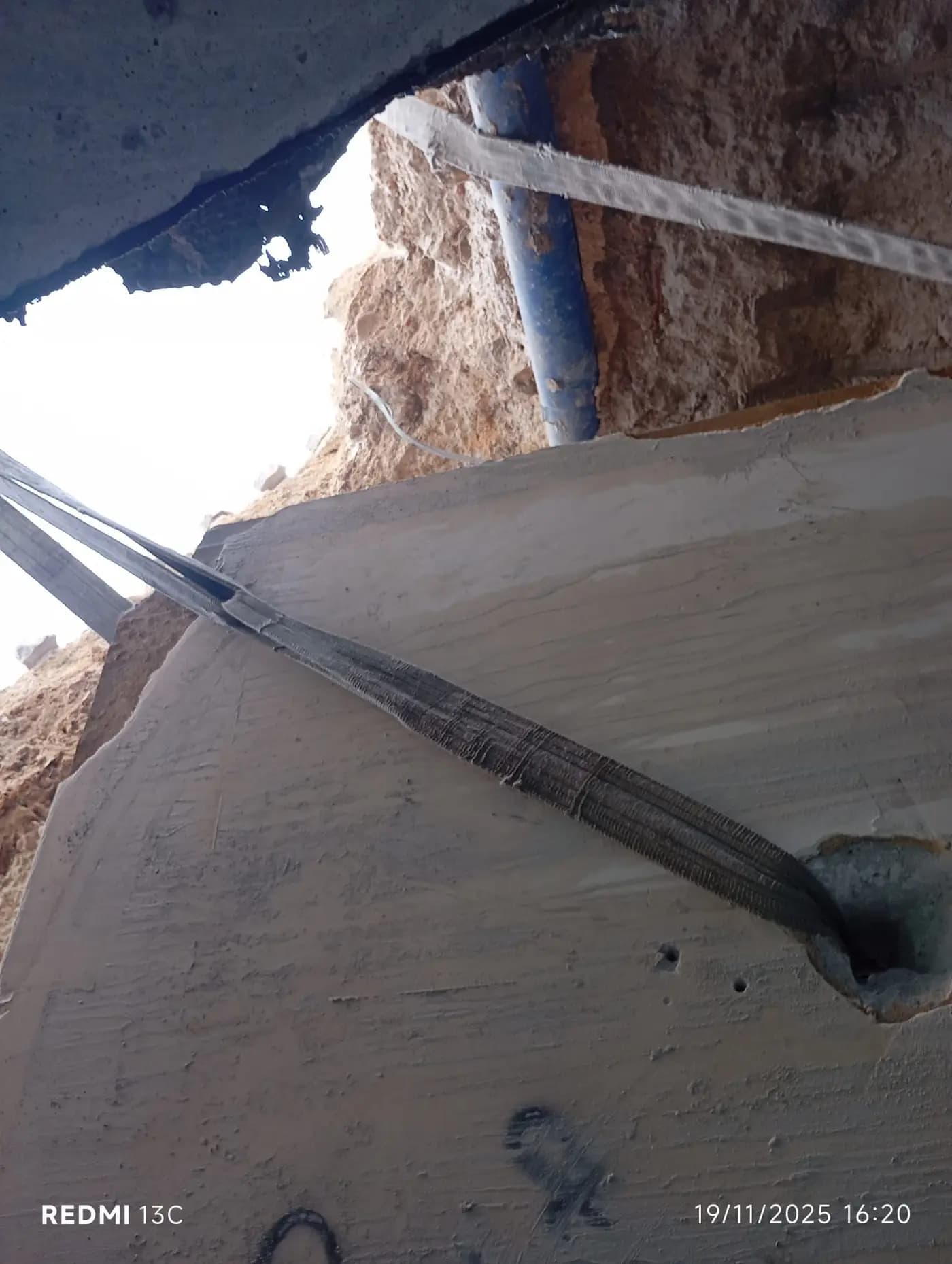 Kesilmiş beton panelin karot deliğinden geçirilmiş sapanla askıya alınmış hâli ve arkadaki toprak ile tesisat borusu