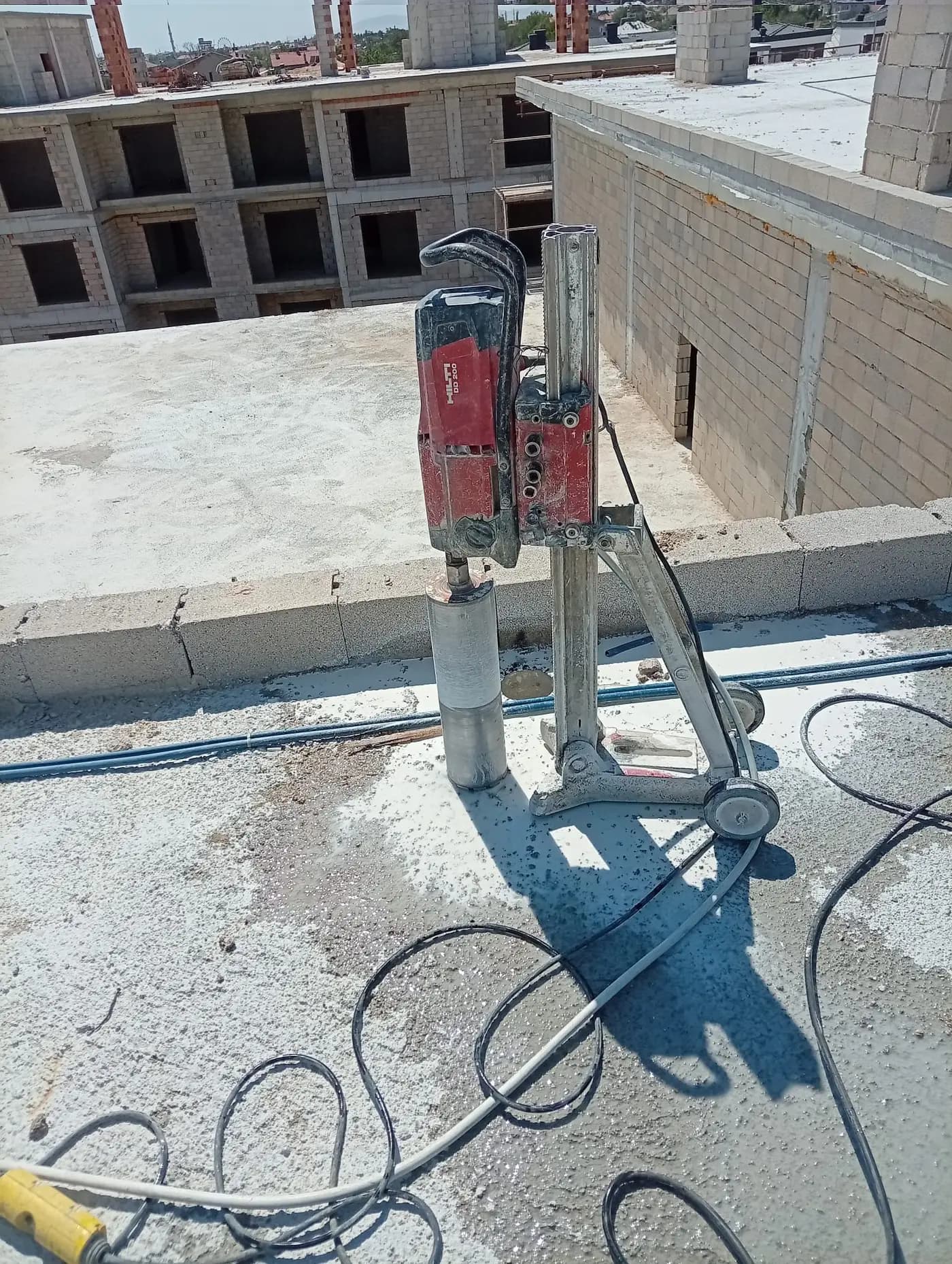 Parapet duvarının önünde döşemeye kurulmuş Hilti DD 200 karot makinesi, elmas karot ucu ve yanından geçen mavi tesisat boruları