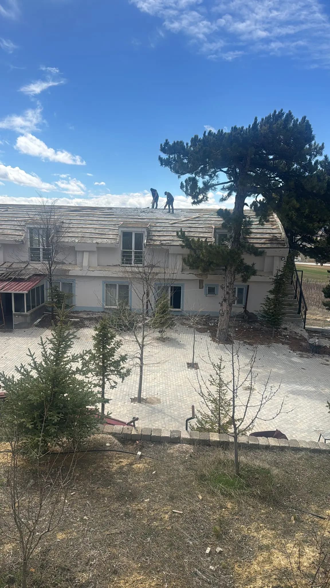 İki katlı tesis binasının kırma çatısı üzerinde kaplamayı söken iki işçi ve kilit taşı kaplı avlu
