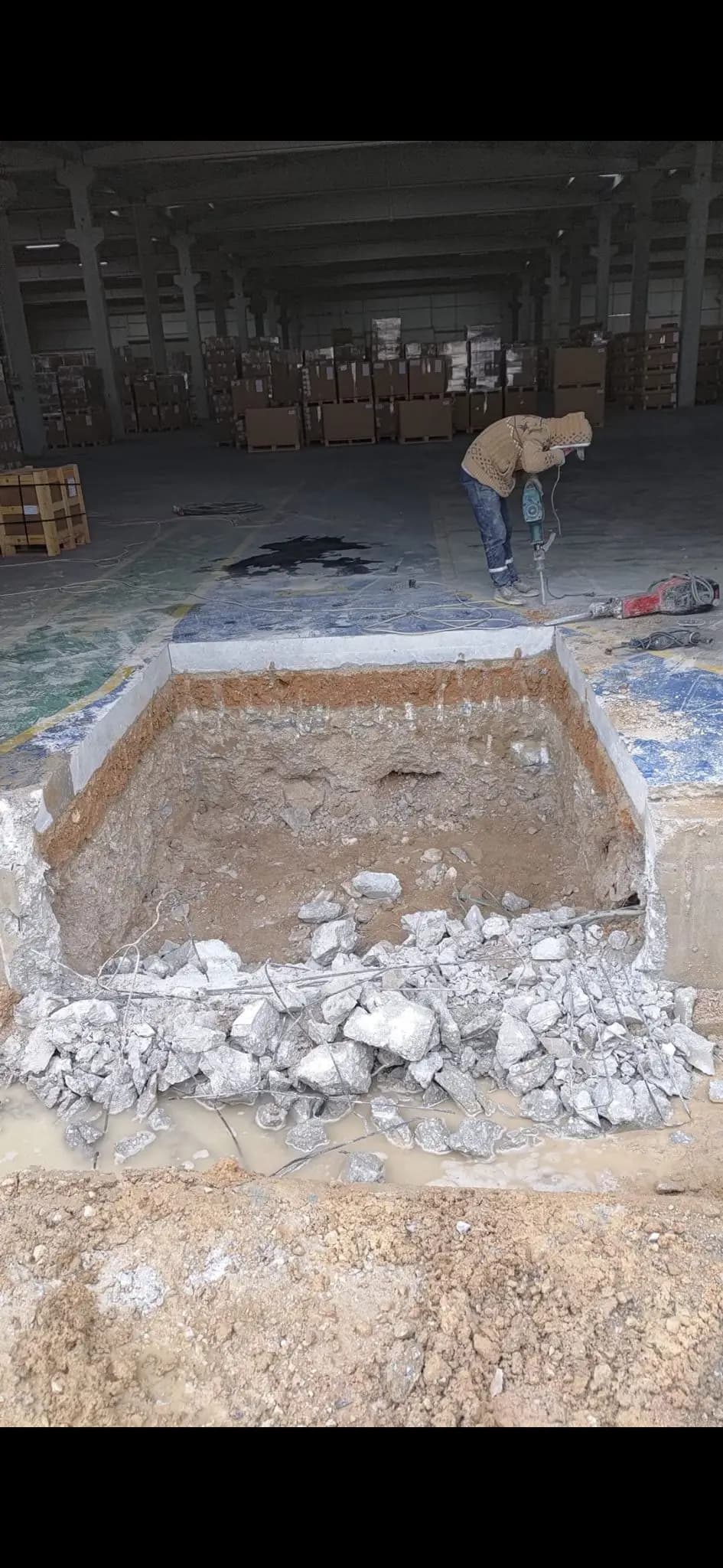 Paletli mal istiflenmiş depoda zemin betonu kesilerek toprağa kadar açılmış dikdörtgen çukur ve arkada kırıcıyla çalışan işçi