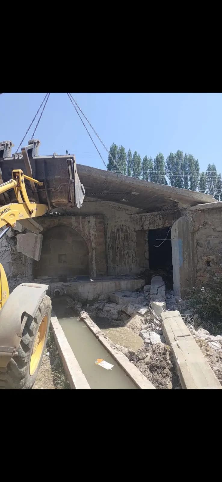 Taş duvarlı yapının betonarme saçağını kovayla zorlayan sarı lastikli yükleyici ve yapının önünden geçen sulama kanalı