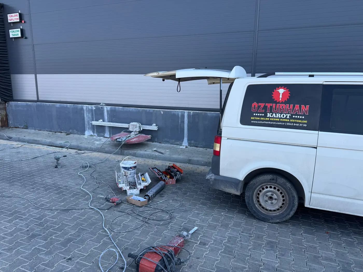Tesis yükleme rampası önünde beton bordüre kurulmuş raylı testere, kilit taşı üzerine serilmiş ekipman ve Özturhan Karot servis aracı